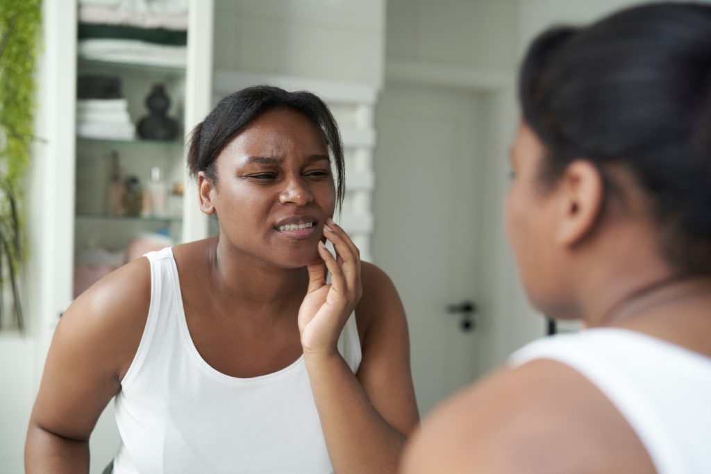 young woman looking in mirror holding her jaw in pain, signs of bruxism